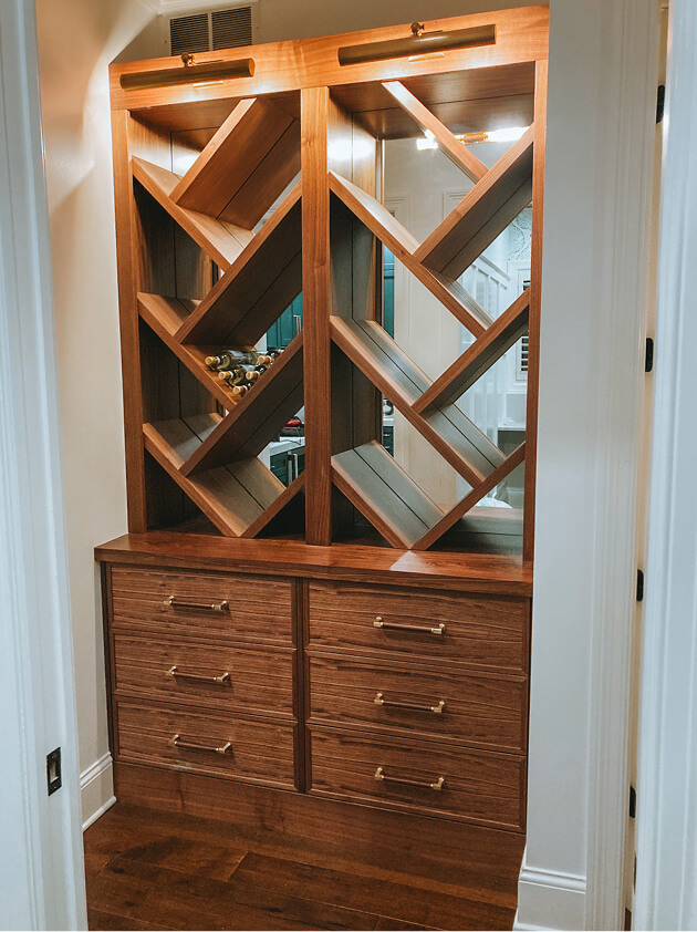 Built-in wooden wine rack with diagonal shelves holds a few wine bottles above a set of four drawers with brass handles, set in a recessed alcove with white walls.