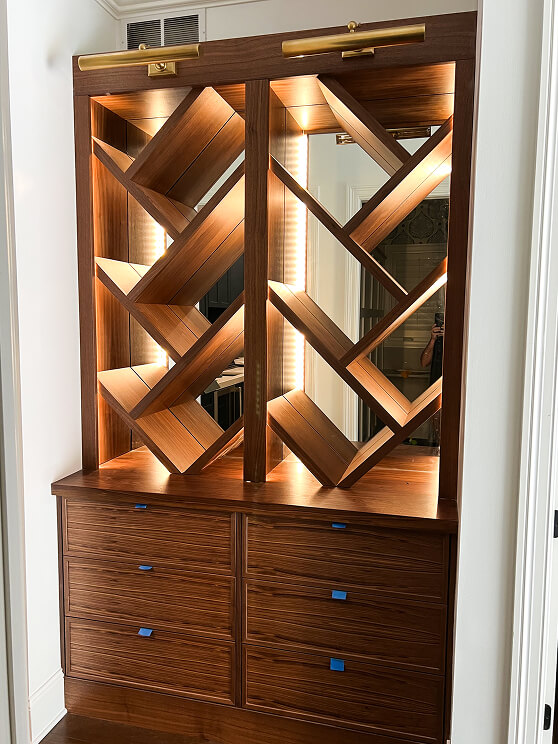 A wooden wine rack with diagonal shelves and built-in lighting is mounted above a set of drawers with blue painter’s tape tabs, situated in a white-walled room.