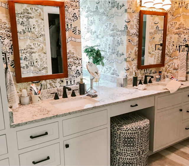 Double bathroom vanity with two mirrors, a patterned stool, and decorative wallpaper. The countertop holds toiletries and a small potted plant.