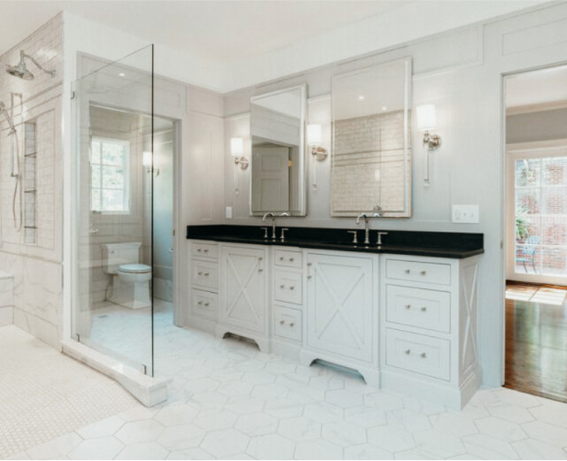 Modern bathroom with double vanity, black countertop, large mirrors, glass-enclosed shower, and white hexagonal tile floor. Toilet is visible in a separate alcove.