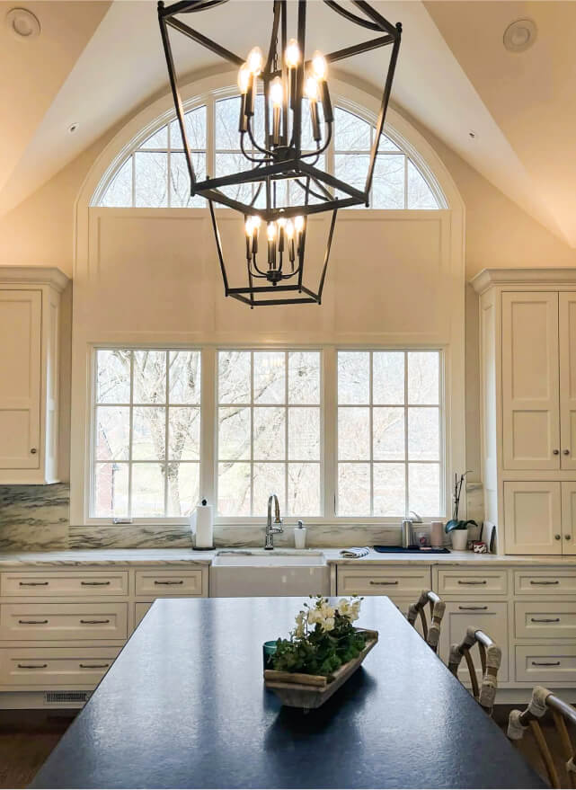 Modern kitchen with cream cabinets, farmhouse sink, marble countertops, large arched window, and geometric pendant lights above a dark island with a small plant centerpiece.