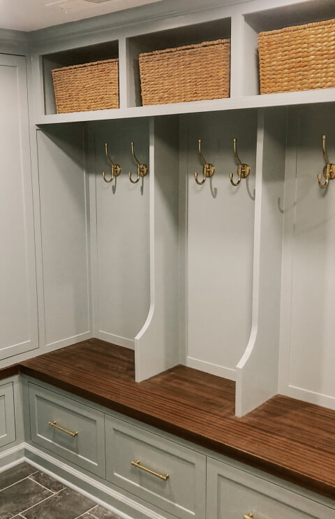 Built-in mudroom storage with three wooden cubbies, each with brass hooks, a wood bench, drawers below, and woven baskets on the top shelf.