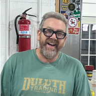 A man with glasses and a beard, wearing a green Duluth Trading t-shirt, stands smiling indoors near a fire extinguisher and a metal cabinet with stickers.