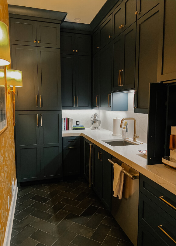 A small kitchen with dark green cabinets, white countertops, a sink, books, and appliances. Herringbone tile flooring and gold hardware are visible.