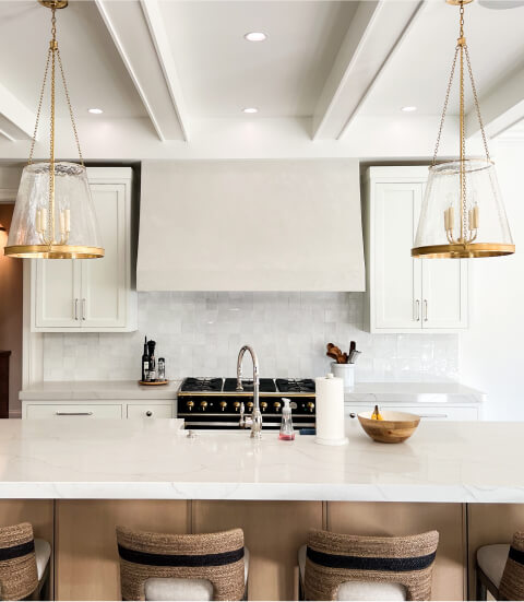 Modern kitchen with a large island, gold pendant lights, white cabinetry, a stainless steel sink, and a stove with a range hood against a white tile backsplash.