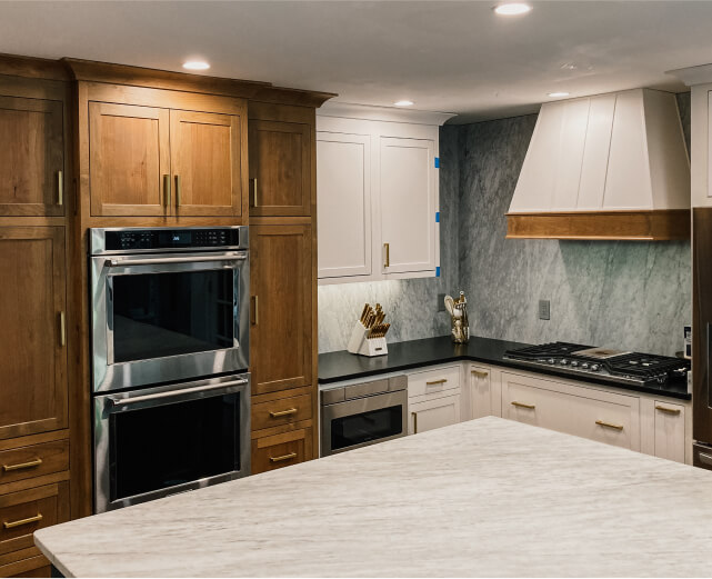 Modern kitchen with wooden and white cabinets, stainless steel double oven, marble countertops, and a gas cooktop with a white range hood.