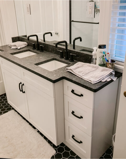 A modern bathroom with a double sink vanity, black faucets, white cabinets, folded towels, soap dispenser, and toiletries on a dark countertop.
