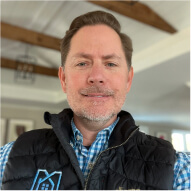 A man with short brown hair and a trimmed beard is wearing a blue checked shirt and a black vest, standing indoors with wooden ceiling beams in the background.