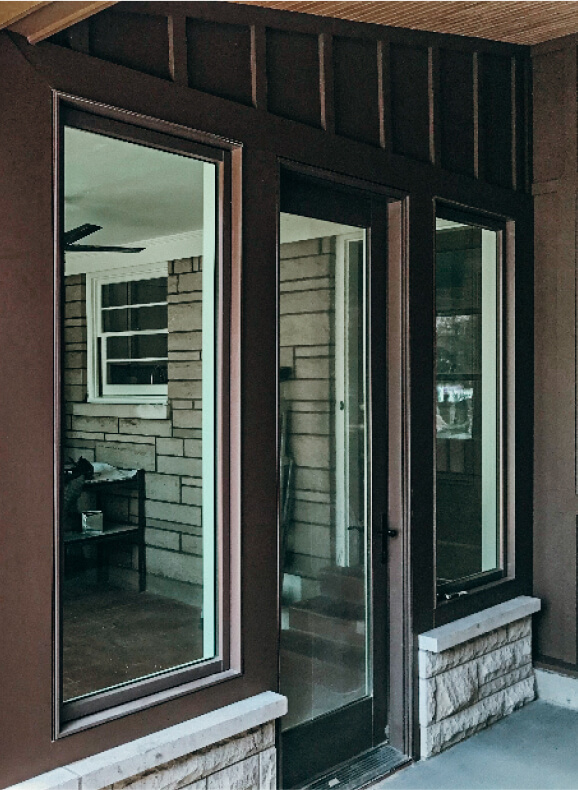 Brown-framed glass door and windows on a porch, reflecting an outdoor scene. An interior room with brick walls and furniture is visible through the glass.