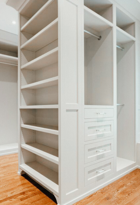 White built-in closet with open shelves, three drawers, hanging rods, and hardwood flooring. The closet is empty and well-lit.