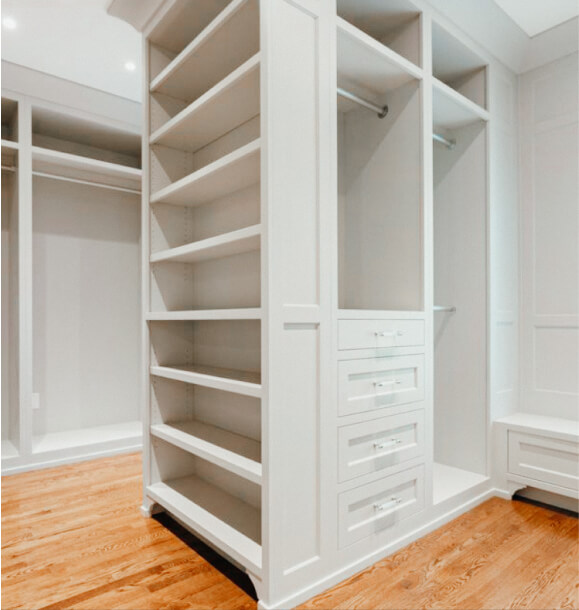 Spacious walk-in closet with built-in white shelves, drawers, and hanging rods on a wooden floor.