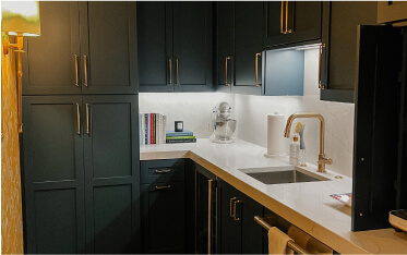 A modern kitchen features dark cabinets, a white countertop, a sink with a gold faucet, under-cabinet lighting, books, and a stand mixer on the counter.