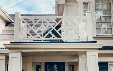 A two-story house with white siding and a decorative white railing above the porch; a Halloween mask is visible in a window by the entrance.