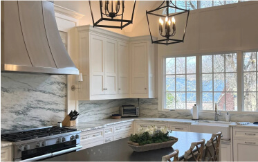 Modern kitchen with white cabinets, marble backsplash, stainless steel stove, large windows, and black pendant lights over a dark countertop with stools.