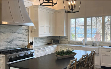 Modern kitchen with white cabinets, marble backsplash, large stainless steel stove hood, black island countertop, and windows overlooking trees and a blue roof.