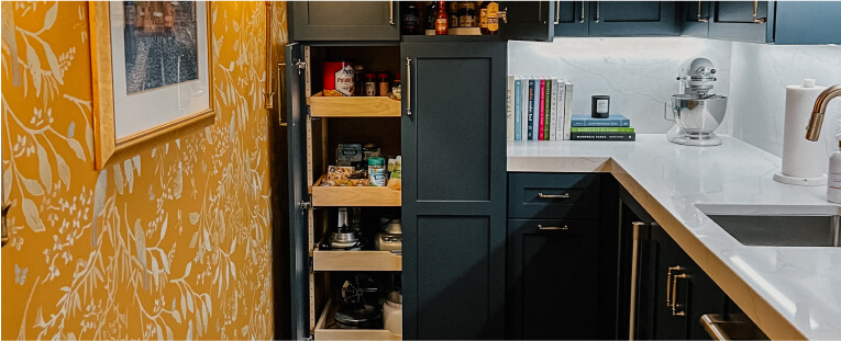 A kitchen with dark cabinets, pull-out pantry shelves holding various items, a mixer, books on the counter, and a yellow floral-patterned wall on the left.