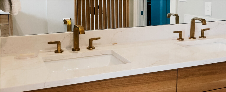 Double bathroom sink with a white marble countertop, brass faucets, wooden cabinets below, and a large mirror above.