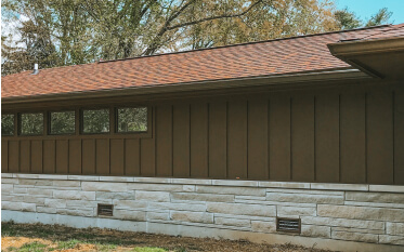 A brown house exterior with vertical siding, a row of small windows, and a lower section of light-colored stone blocks.