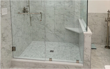 Glass-enclosed shower with marble tile walls and floor, a corner bench, wall-mounted grab bar, and metal fixtures.
