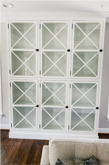 A white built-in cabinet with glass doors featuring a crisscross design, standing against a light-colored wall with a wood floor in the room.