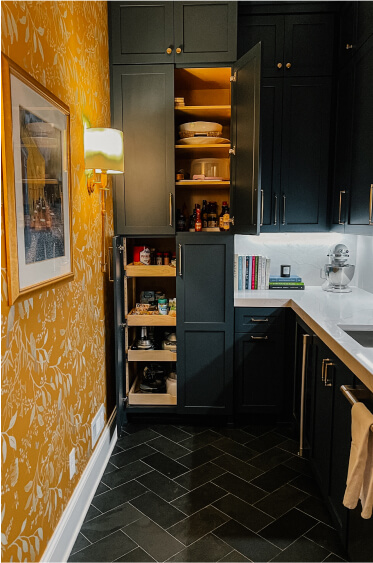 A modern kitchen corner with dark cabinets, pull-out pantry shelves, and upper cabinets open to reveal dishes; yellow wallpaper with a floral pattern on one wall.
