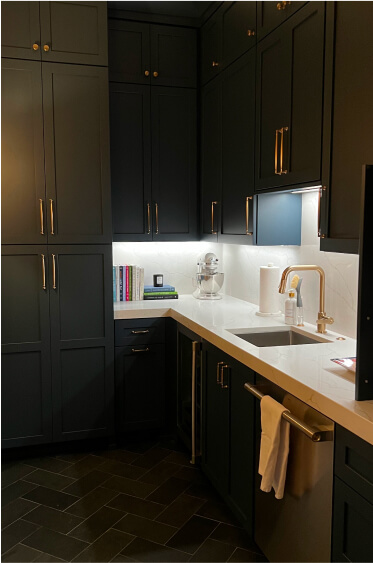 Modern kitchen corner with dark cabinets, white countertops, a sink, built-in shelves with books, a stand mixer, paper towel roll, and under-cabinet lighting.