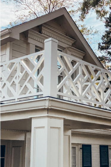 A second-story balcony with a white geometric railing on a beige house, featuring decorative trim and a gabled roof.