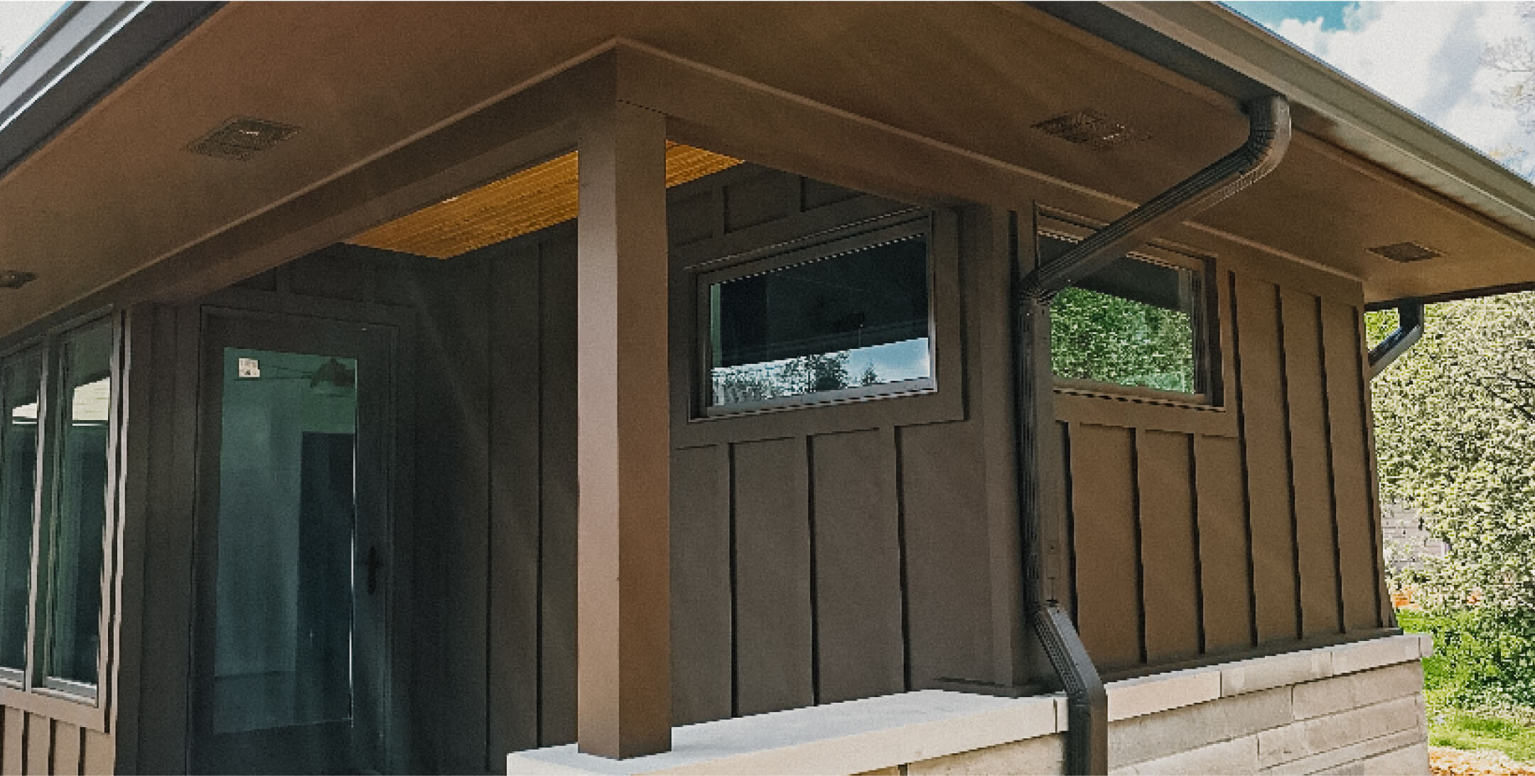Modern house exterior with dark brown siding, black window frames, and a black rain gutter, set against a backdrop of green trees and blue sky.