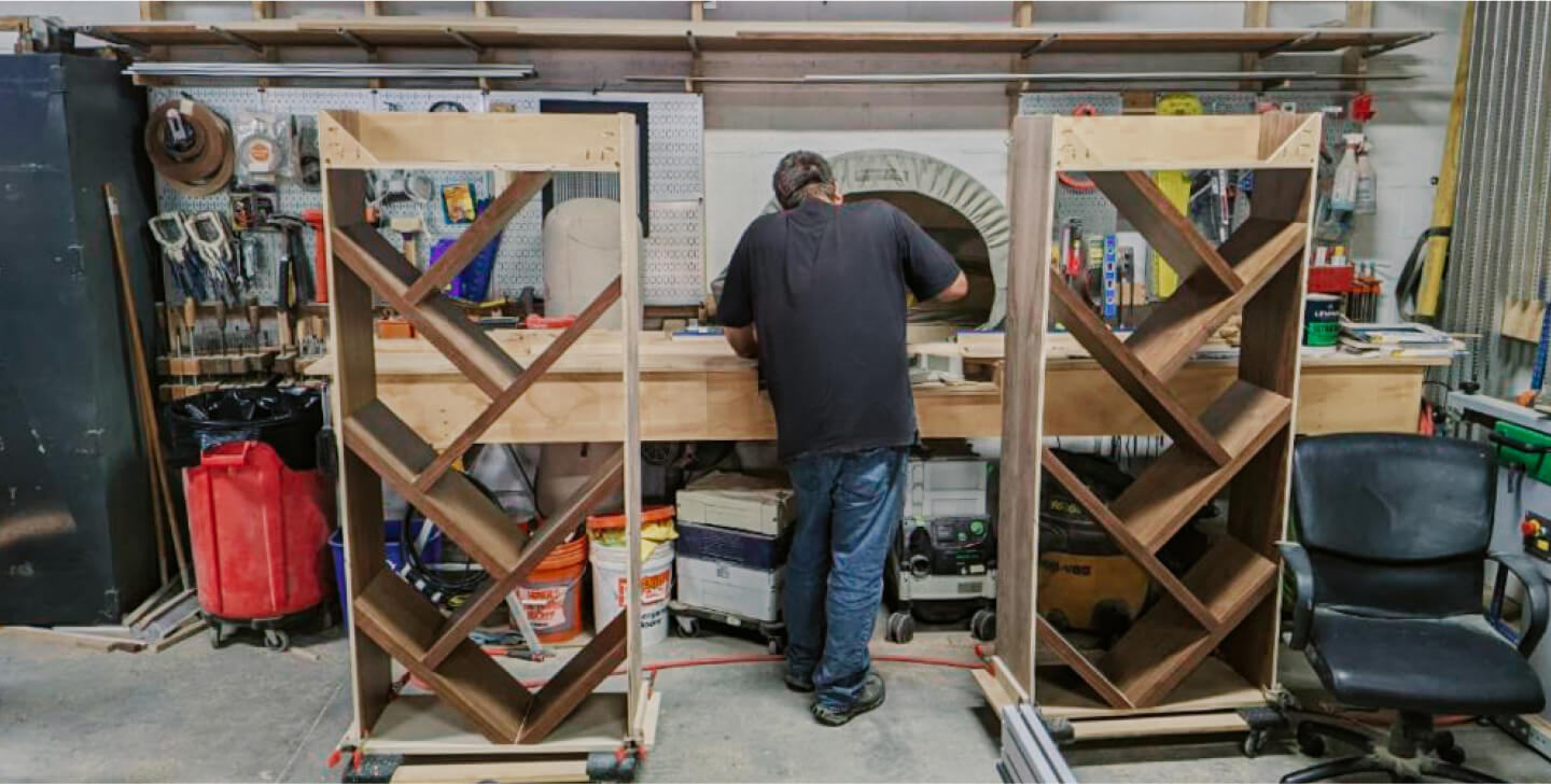 A person works at a bench in a workshop, surrounded by tools and equipment, with two unfinished wooden shelving units in the foreground.
