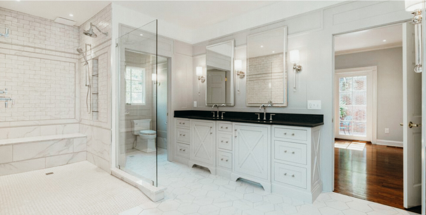 Modern bathroom with double vanity, large mirrors, glass shower enclosure, white cabinetry, and hexagonal floor tiles. Doorway leads to a room with hardwood flooring.