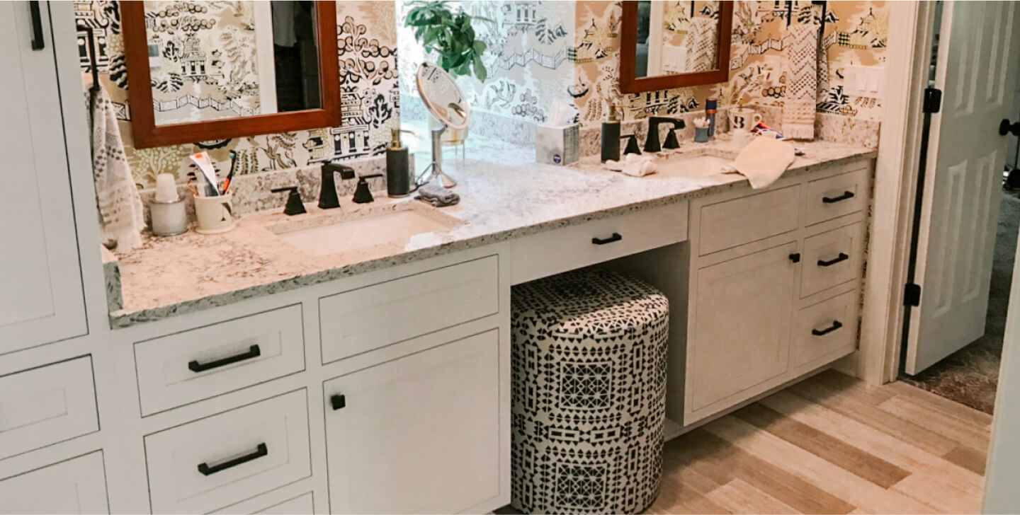 Double bathroom vanity with two sinks, dark fixtures, large mirrors, patterned stool, white cabinets, and decorative wallpaper. Towels and toiletries are on the countertop.